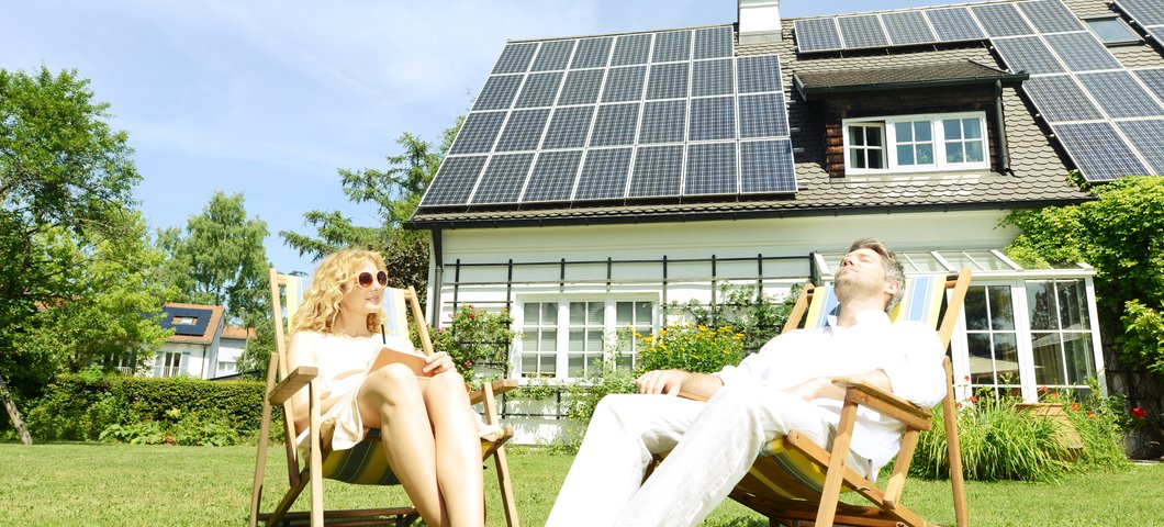 Zwei Personen im Liegestuhl bei Sonnenschein im Garten sitzend vor Haus mit Photovoltaik-Anlage auf dem Dach